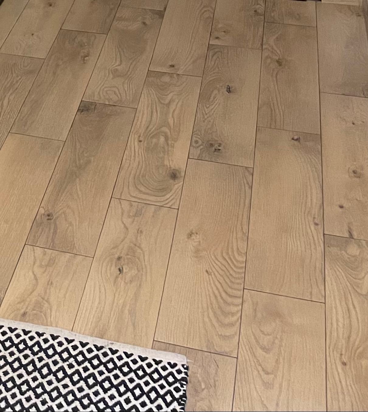 Ułożone panele podłogowe w odcieniach jasnego drewna, widzoczne z góry, z fragmentem czarno-białego dywanika w lewym dolnym rogu.