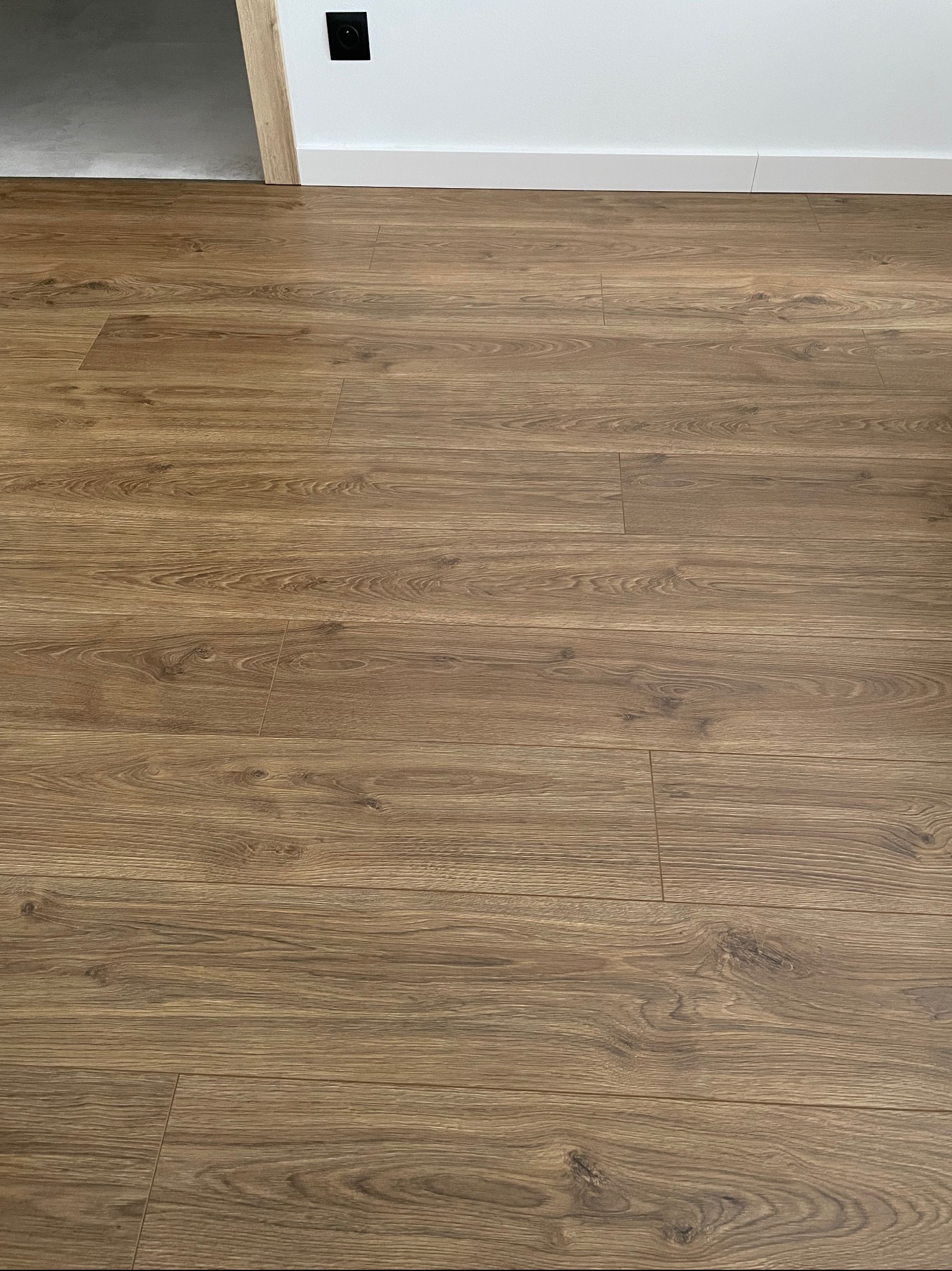 Ułożone panele podłogowe w kolorze jasnego dębu, widoczne delikatne wzory słojów, listwa przypodłogowa i fragment ościeżnicy drzwiowej w tle.