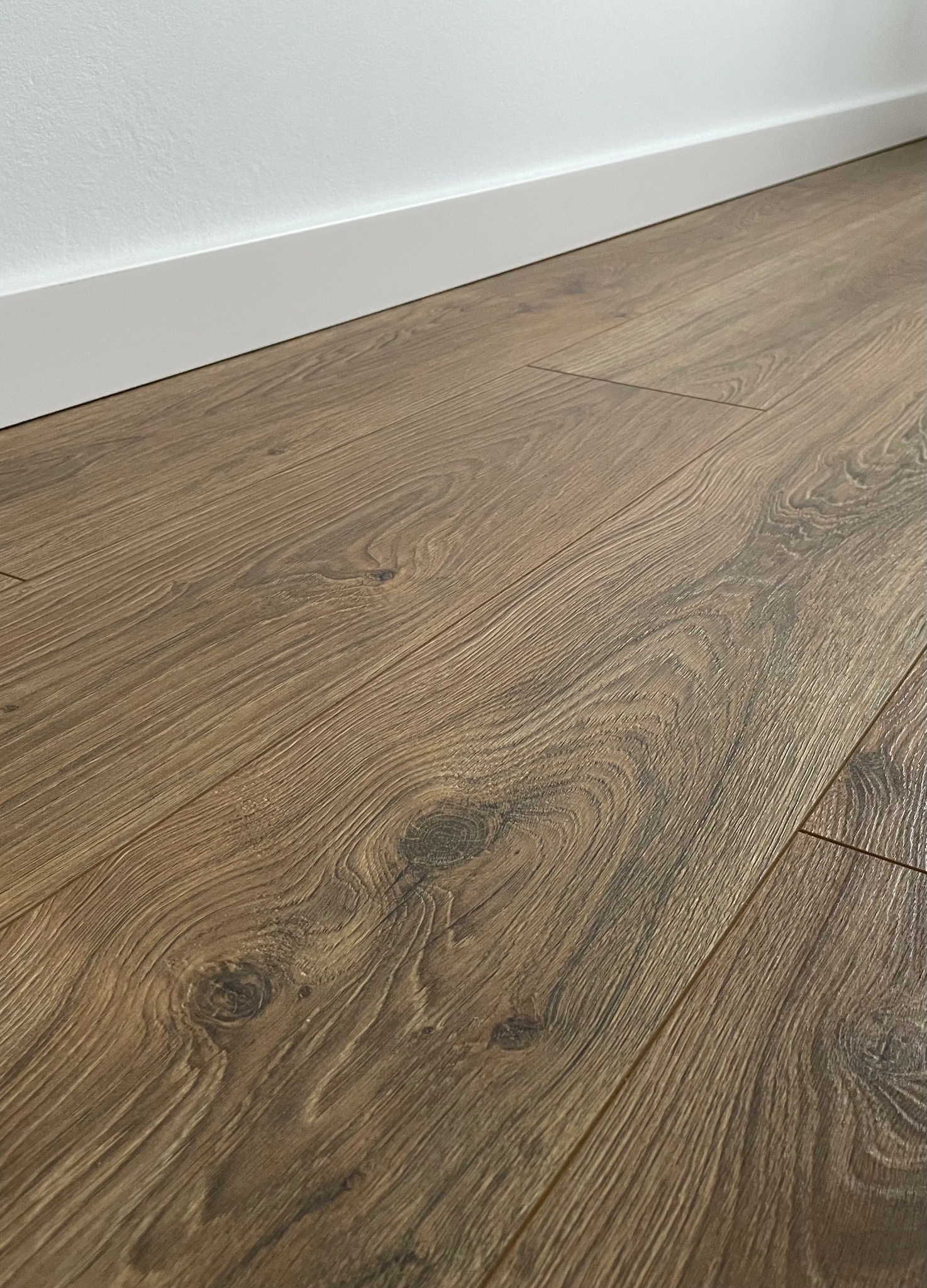 Ułożone panele podłogowe w odcieniach brązu, z widoczną strukturą drewna i białymi listwami przypodłogowymi przy ścianie.