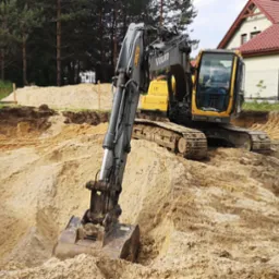 Żółta koparka Volvo na terenie budowy, wykop pod fundamenty domu jednorodzinnego, widoczne drewniane słupki wyznaczające granice wykopu.