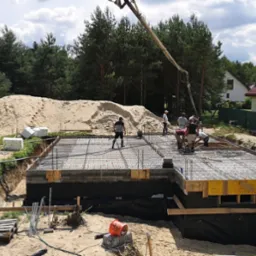 Wylewanie fundamentów domu jednorodzinnego; betoniarka z pompą podaje beton na zbrojenie; widoczni pracownicy budowlani i otoczenie: las, domy.