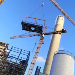 Montaż elementu konstrukcyjnego za pomocą żurawia wieżowego na placu budowy. W tle komin i budynek w trakcie wznoszenia. Ujęcie z dołu, niebo błękitne.