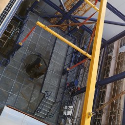 Działalność jednoosobowa - Montaż konstrukcji stalowej w Pysznicy z użyciem dźwigu. Widok z góry na elementy niebieskie i żółte oraz siatkową podłogę.