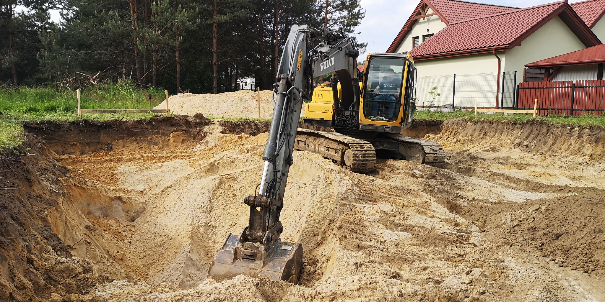 Żółta koparka Volvo na terenie budowy, wykop pod fundamenty domu jednorodzinnego, widoczne drewniane słupki wyznaczające granice wykopu.