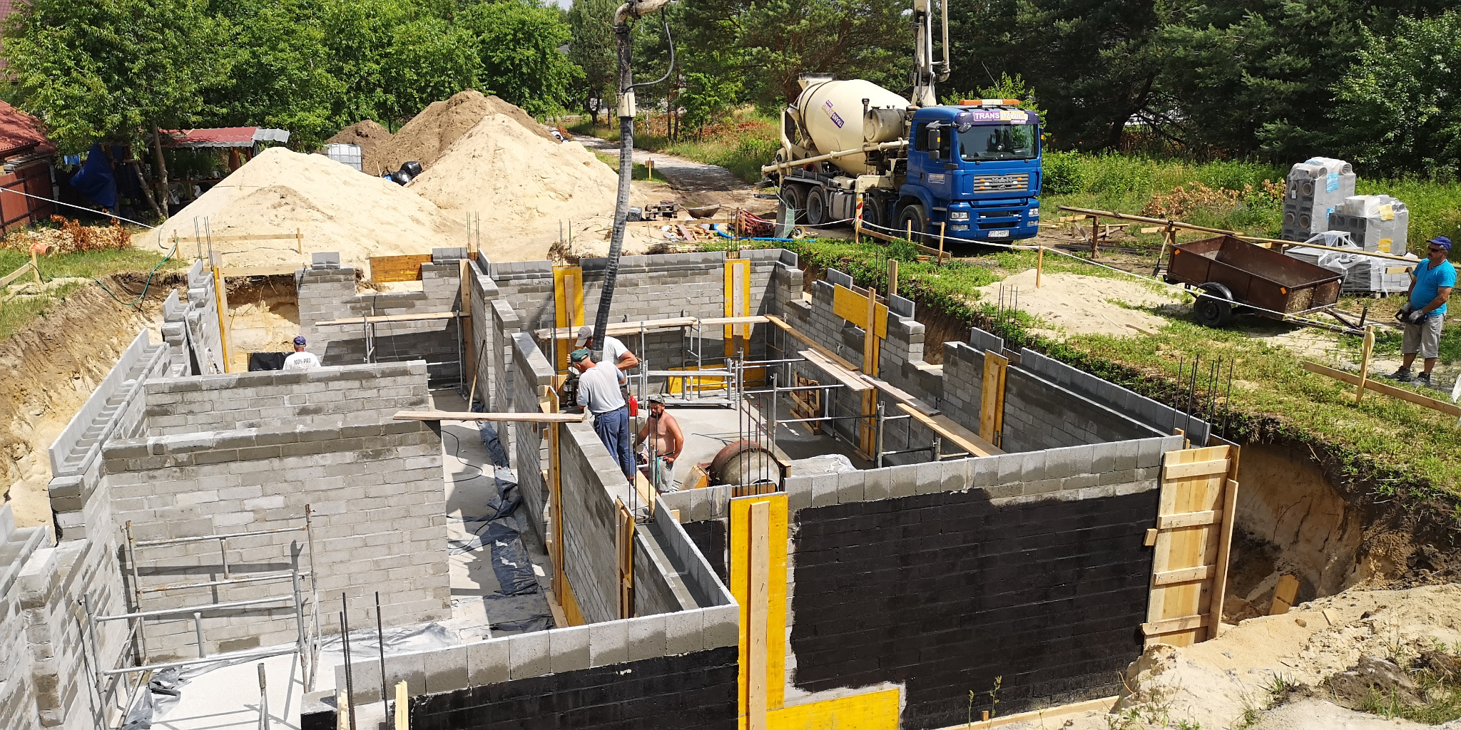 Wykop pod fundamenty domu z częściowo wylanymi ścianami z bloczków betonowych, zabezpieczonymi czarną izolacją; na budowie obecni robotnicy i betonowóz z pompą do betonu.