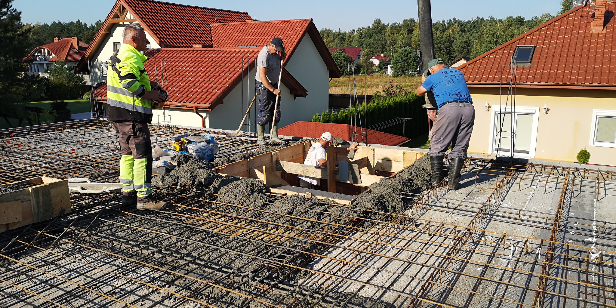 Wylewanie betonu na zbrojenie dachu domu jednorodzinnego; widoczni pracownicy w roboczych ubraniach i kaloszach, domy w tle.