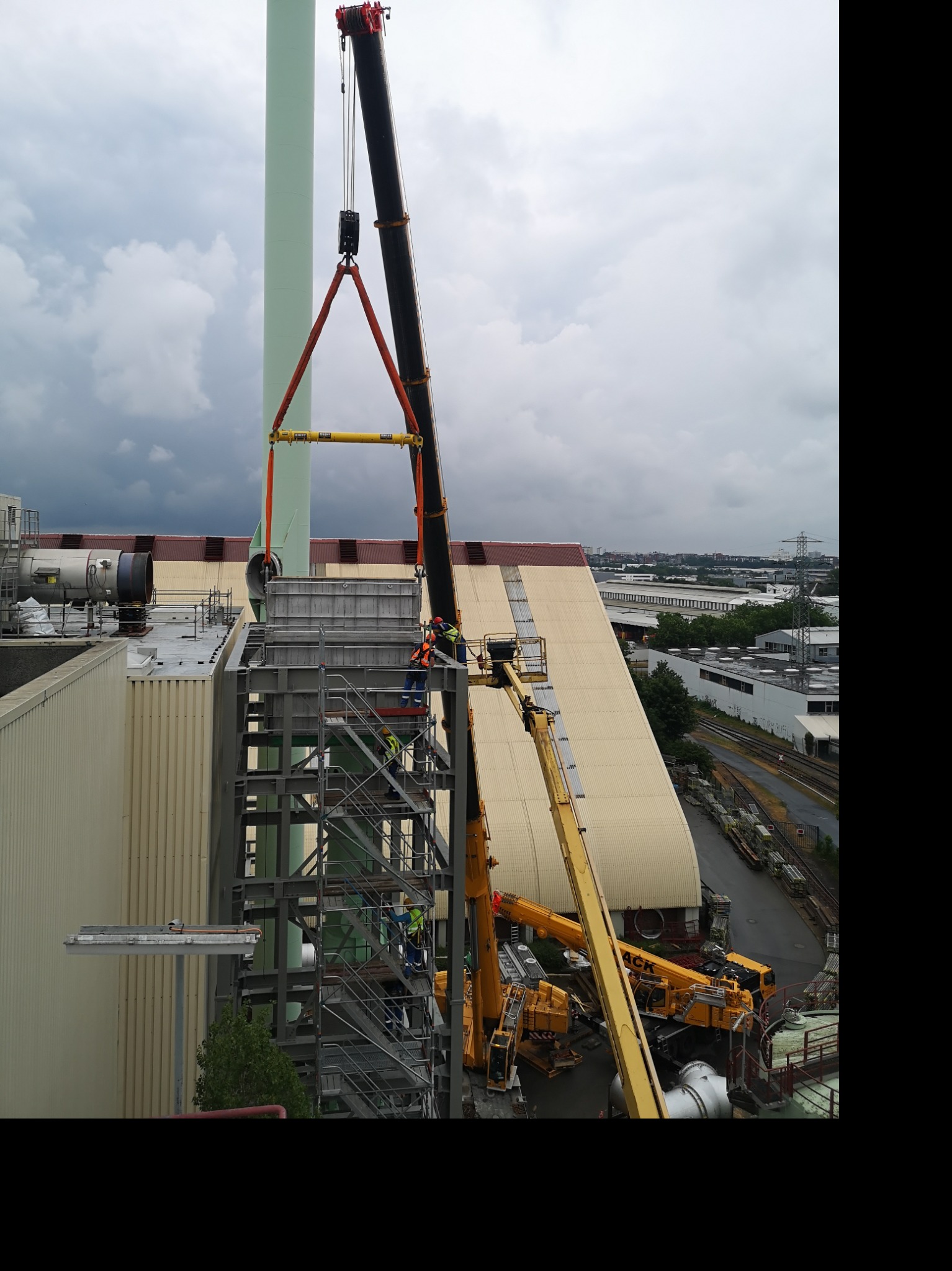 Montaż stalowej konstrukcji na dachu budynku przemysłowego przy użyciu dźwigu. Pracownicy w kaskach nadzorują proces. Widoczne inne budynki i pochmurne niebo.