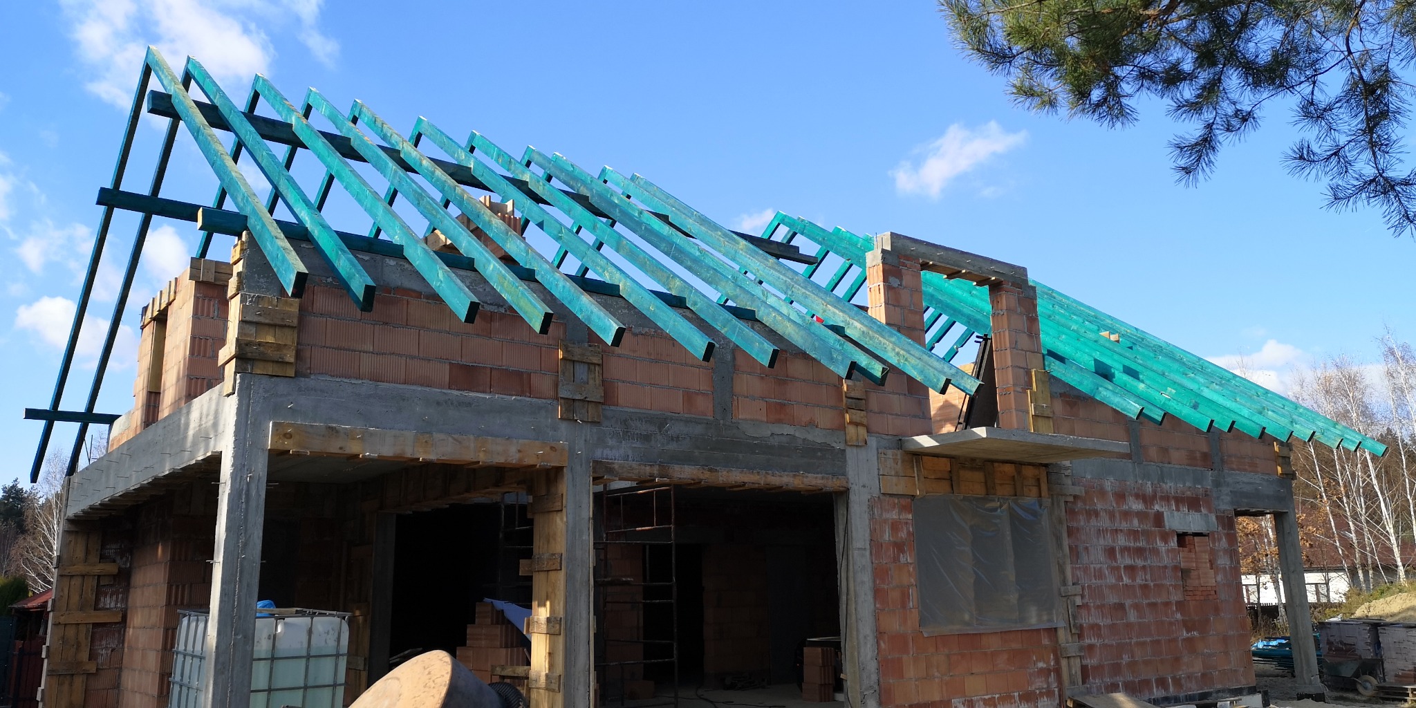 Budowa domu z czerwonej cegły: widok na konstrukcję dachu z zielonymi elementami na tle błękitnego nieba. Widoczne rusztowania i fragmenty otoczenia.