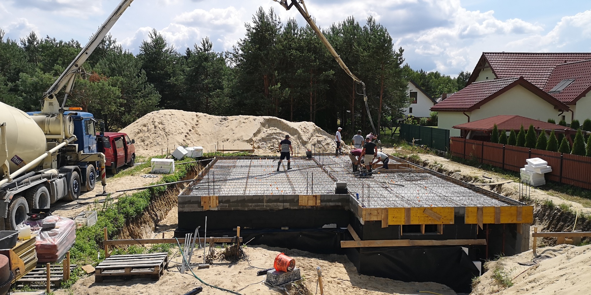 Wylewanie fundamentów domu jednorodzinnego; betoniarka z pompą podaje beton na zbrojenie; widoczni pracownicy budowlani i otoczenie: las, domy.