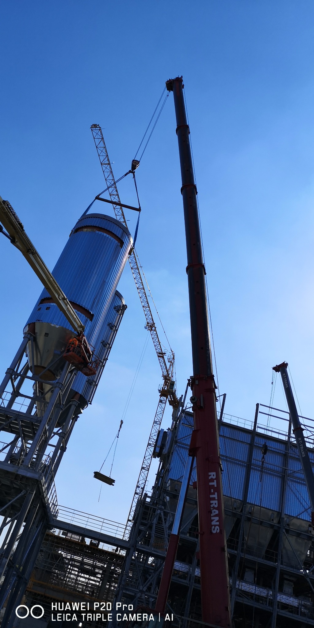 Montaż stalowych silosów na budowie hali przemysłowej przy użyciu dźwigów. Widok z dołu na konstrukcje i błękitne niebo.