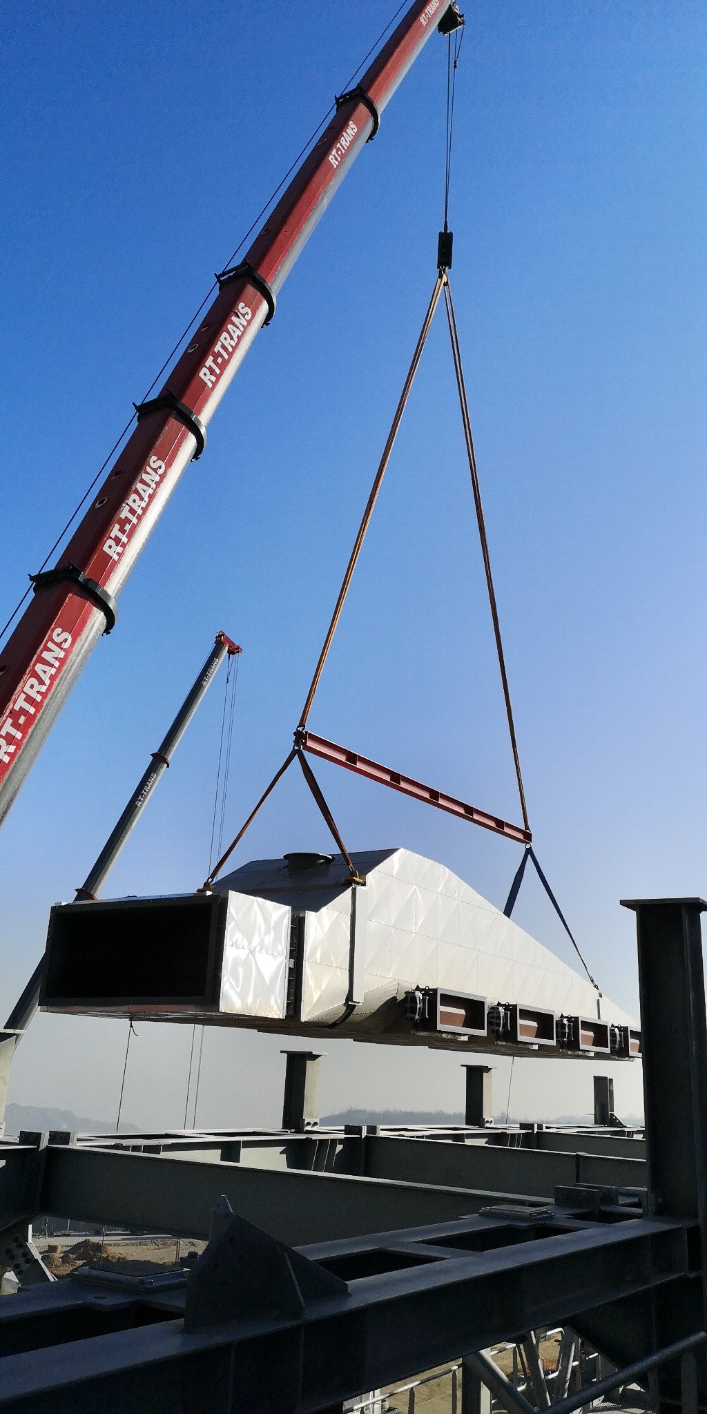 Montaż elementu hali stalowej za pomocą dźwigu RT-TRANS na tle błękitnego nieba. Widoczna konstrukcja stalowa i odległe wzgórza.