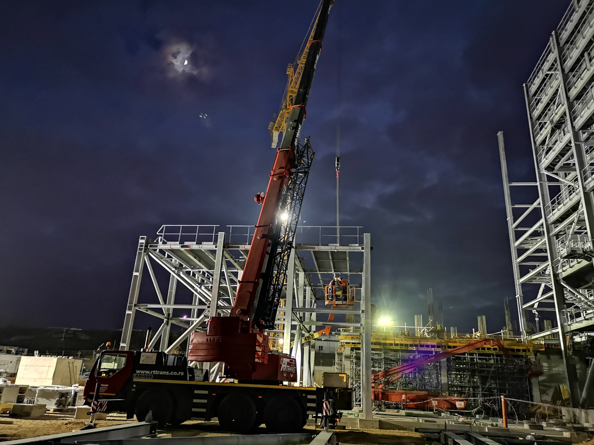 Nocna budowa hali z konstrukcji stalowych, widoczny żuraw Liebherr i pracownicy na podnośniku. W tle księżyc i ciemne niebo.