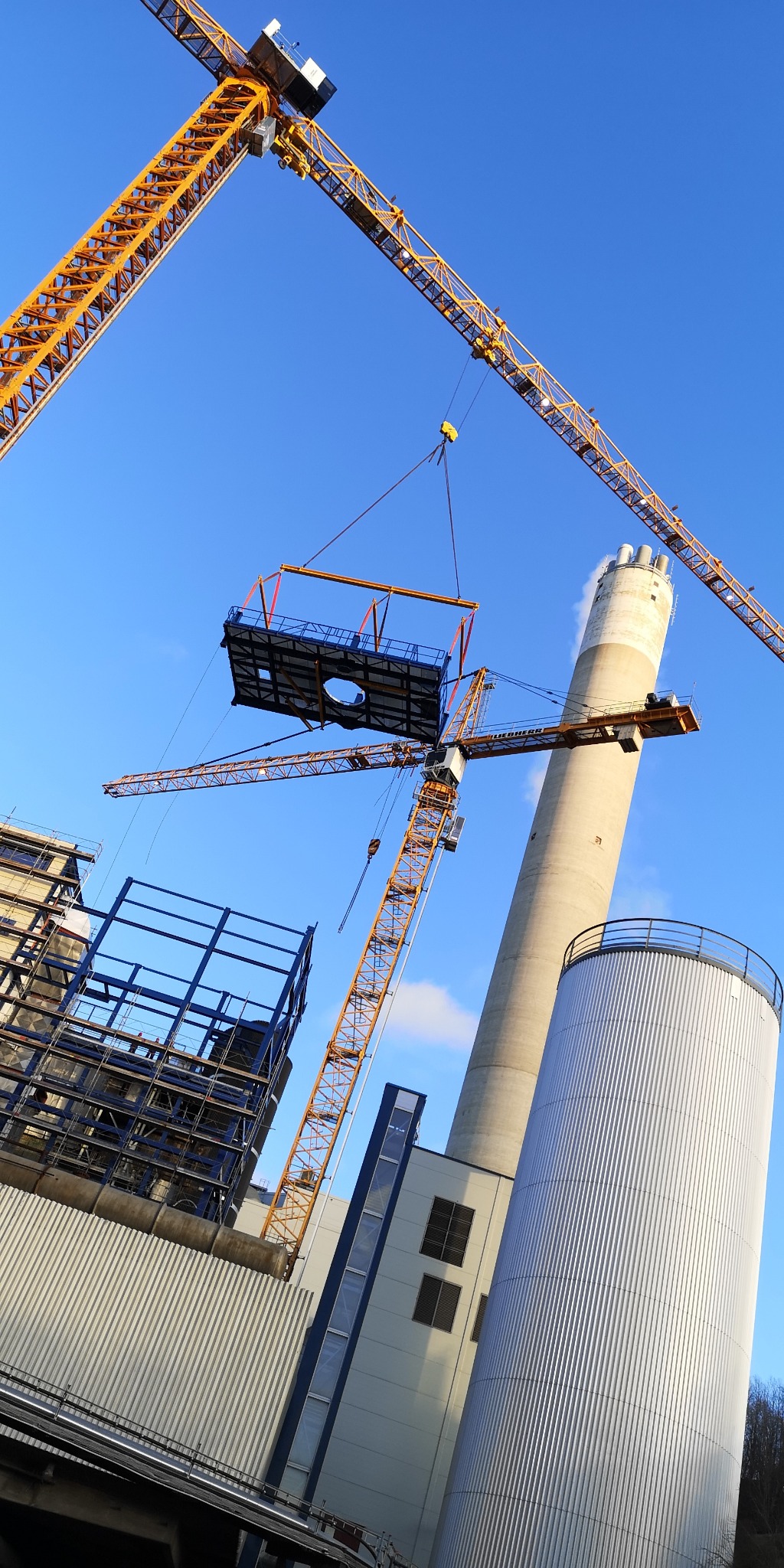 Montaż elementu konstrukcyjnego za pomocą żurawia wieżowego na placu budowy. W tle komin i budynek w trakcie wznoszenia. Ujęcie z dołu, niebo błękitne.