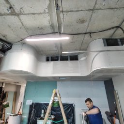 Perfect Wall - Prace wykończeniowe wnętrza: montaż i szpachlowanie płyty karton-gips na suficie z widoczną konstrukcją, oświetleniem i pracownikiem przygotowującym się do pracy, drabina z wiadrem farby w tle.