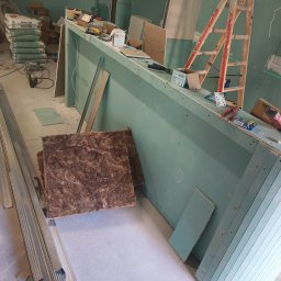 Perfect Wall - Wnętrze w trakcie prac wykończeniowych: widoczne płyty gipsowo-kartonowe na ścianach, metalowe profile, worki z zaprawą murarską, drabina i narzędzia, sugerujące montaż konstrukcji i przygotowanie...