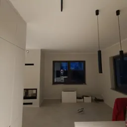 Wnętrze pokoju z minimalistyczną zabudową meblową, kominkiem, trzema wiszącymi lampami i dużym oknem, widoczne ślady po pracach wykończeniowych.