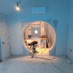 Perfect Wall - Pracownik w trakcie montażu płyty karton gips na okrągłym otworze w ścianie, widoczne narzędzia, szpachlowane ściany i surowy beton na podłodze w pomieszczeniu z klimatyzacją.