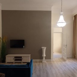 Perfect Wall - Eleganckie wnętrze po odświeżeniu: ściana w odcieniu taupe z telewizorem, dekoracyjny wazon na postumencie, kryształowy żyrandol i parkiet w jasnym odcieniu dębu.