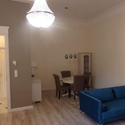 Perfect Wall - Jasne wnętrze: salon z drewnianą podłogą, niebieską sofą, okrągłym stołem z krzesłami, kryształowym żyrandolem i ścianami w odcieniach beżu i szarości.