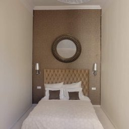 Perfect Wall - Wąska sypialnia z beżową tapetą, pikowanym zagłówkiem, okrągłym lustrem i symetrycznie rozmieszczonymi kinkietami po bokach. Ściany pomalowane na kremowo.