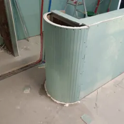 Konstrukcja z płyty karton gips w kształcie zaokrąglonego baru, widoczne narzędzia do cięcia profili i fragmenty płytek na surowym betonowym podłożu, ściany w tle również z płyt karton gips.