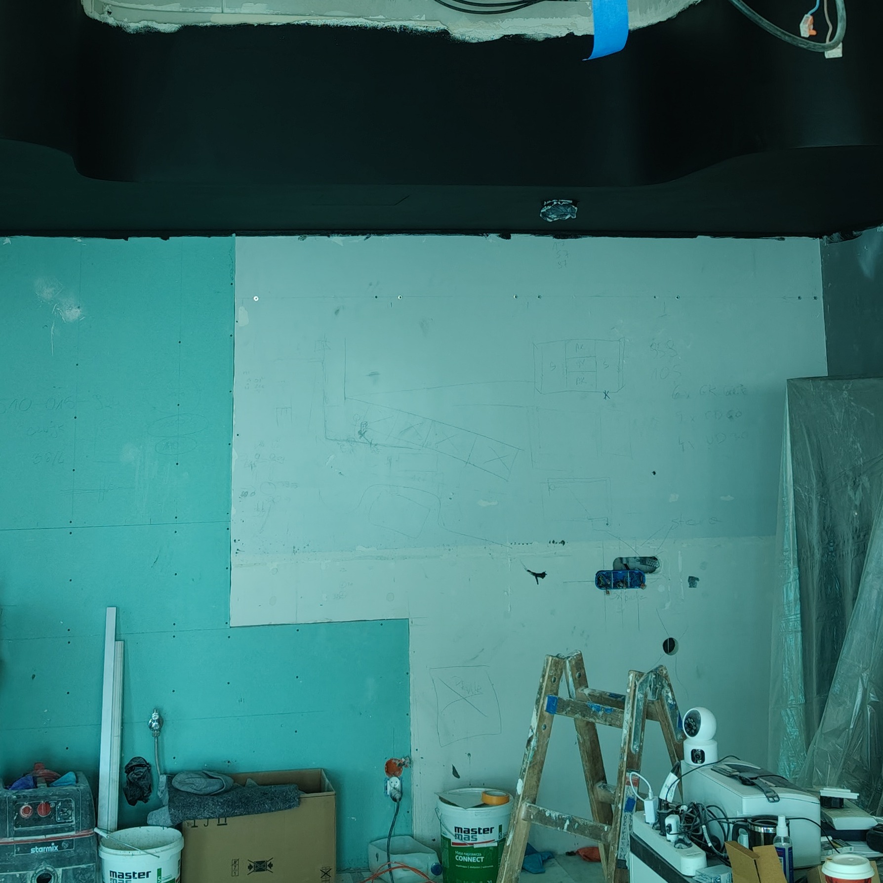 Ściana w trakcie prac wykończeniowych z widocznymi płytami gipsowo-kartonowymi, rysunkami technicznymi i instalacjami elektrycznymi, drabina malarska i narzędzia budowlane w pomieszczeniu z czarnym...