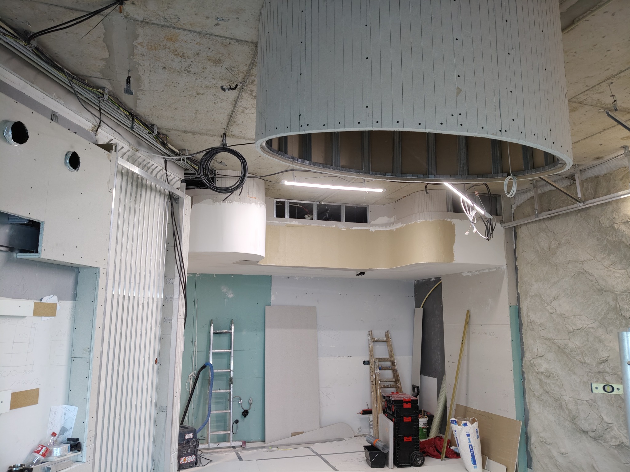 Wnętrze w trakcie prac wykończeniowych: okrągła konstrukcja sufitowa z płyt gipsowo-kartonowych, ściany częściowo pokryte płytami, widoczne drabiny, narzędzia i instalacje elektryczne.