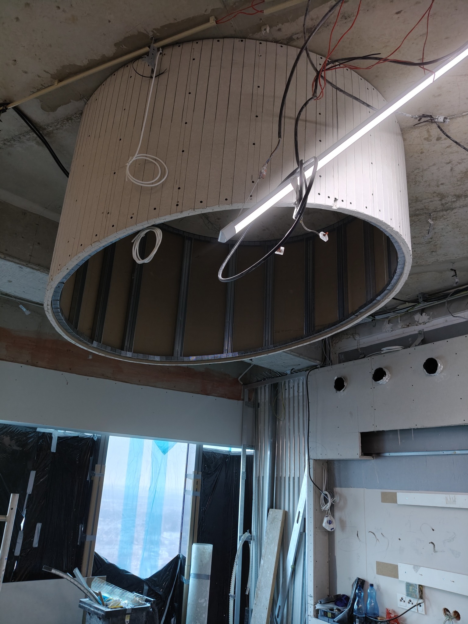 Niedokończona instalacja sufitu podwieszanego z płyty gipsowo-kartonowej o okrągłym kształcie, widoczne metalowe profile konstrukcyjne i przewody elektryczne, surowy betonowy strop w tle.