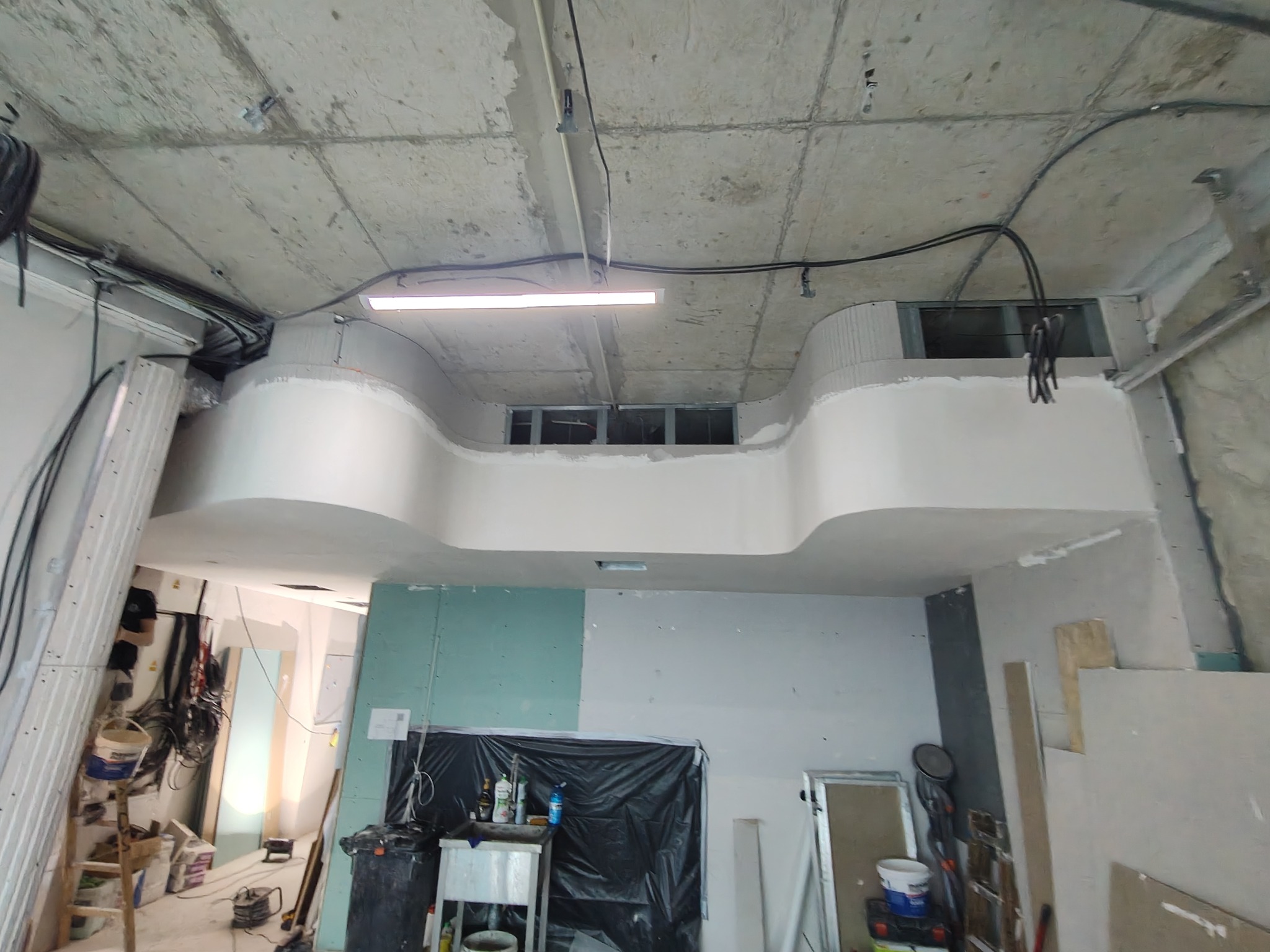 Wnętrze w trakcie remontu z widoczną konstrukcją sufitu z płyt kartonowo-gipsowych, odsłoniętym betonowym stropem, instalacją elektryczną i fragmentami ścian przygotowanych do dalszych prac...