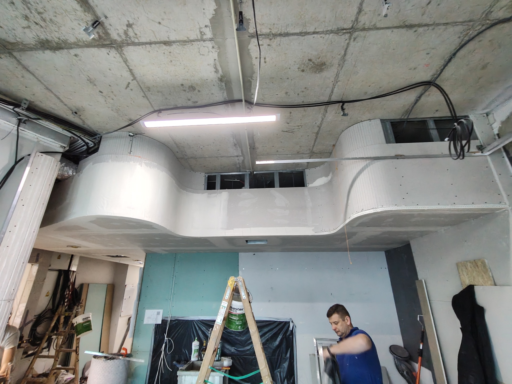 Prace wykończeniowe wnętrza: montaż i szpachlowanie płyty karton-gips na suficie z widoczną konstrukcją, oświetleniem i pracownikiem przygotowującym się do pracy, drabina z wiadrem farby w tle.
