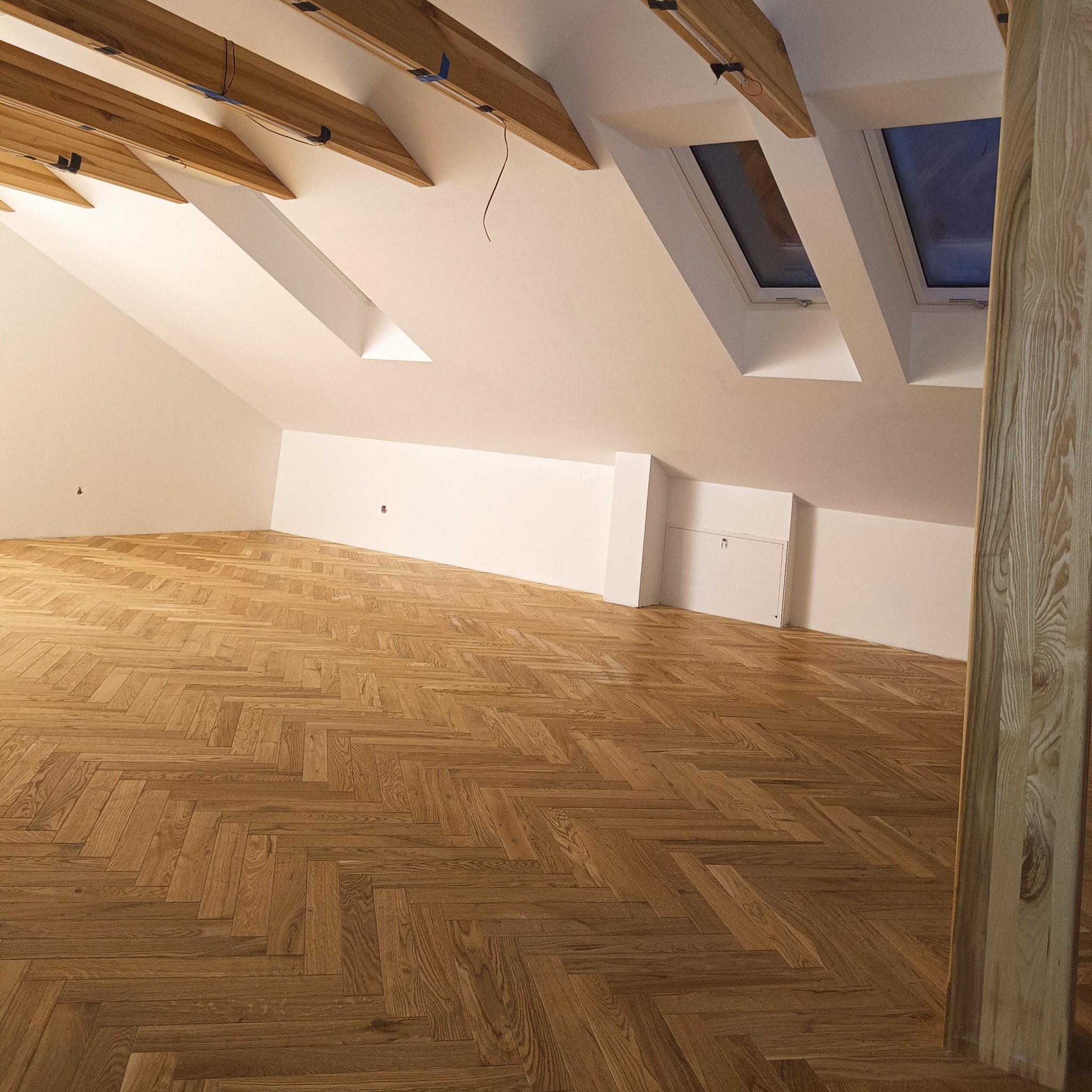 Poddasze z jasną, skośną ścianą, drewnianymi belkami stropowymi, oknami dachowymi i parkietem ułożonym w jodełkę.