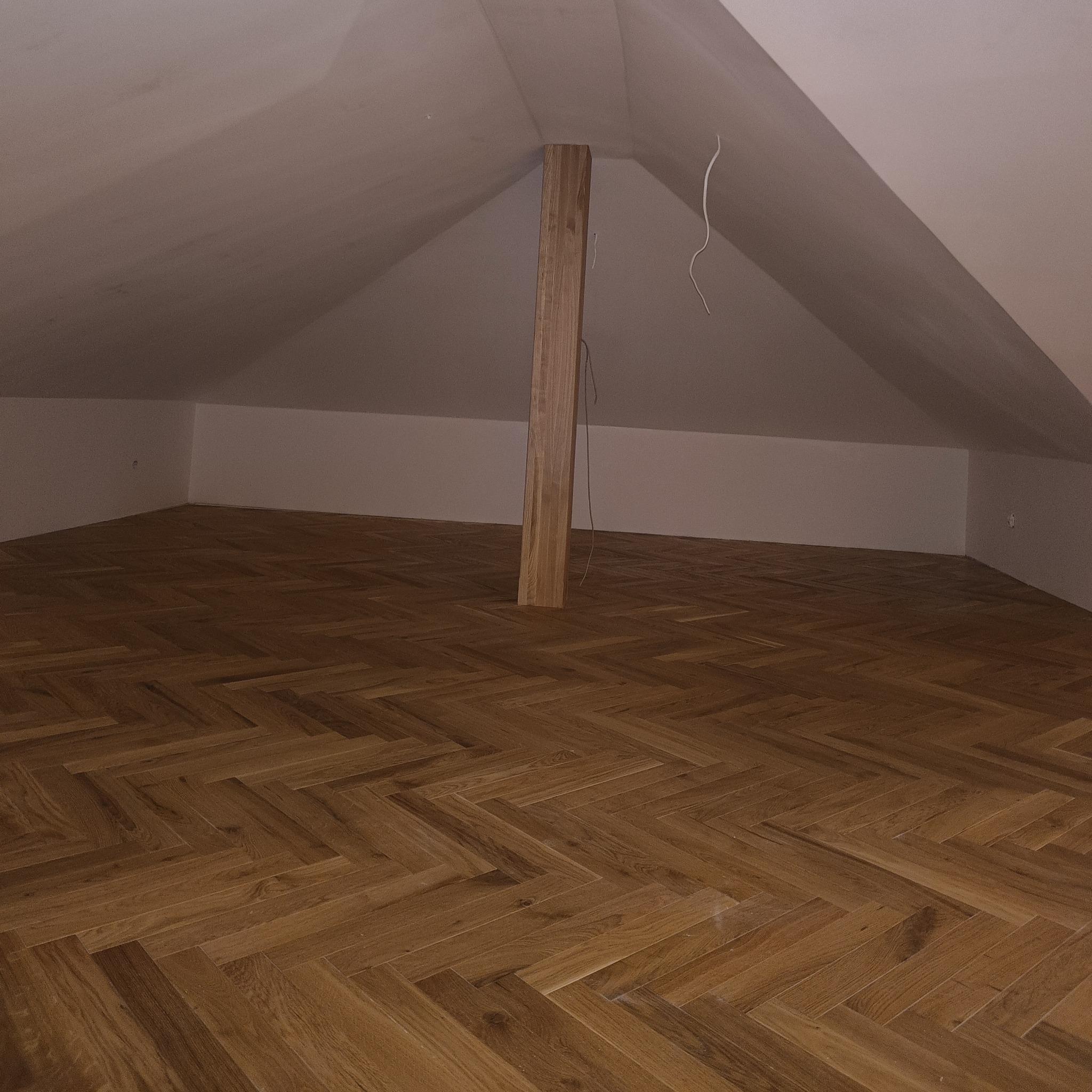 Poddasze z nową podłogą ułożoną w jodełkę, drewniany słup konstrukcyjny na środku, białe ściany i sufit ze skosami.