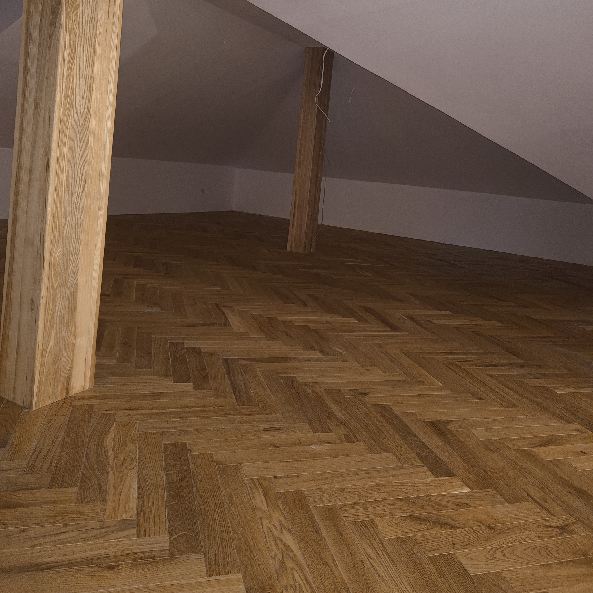 Nowa, jasna podłoga z parkietu ułożonego w jodełkę w pokoju na poddaszu, z widoczną drewnianą kolumną i białymi ścianami.