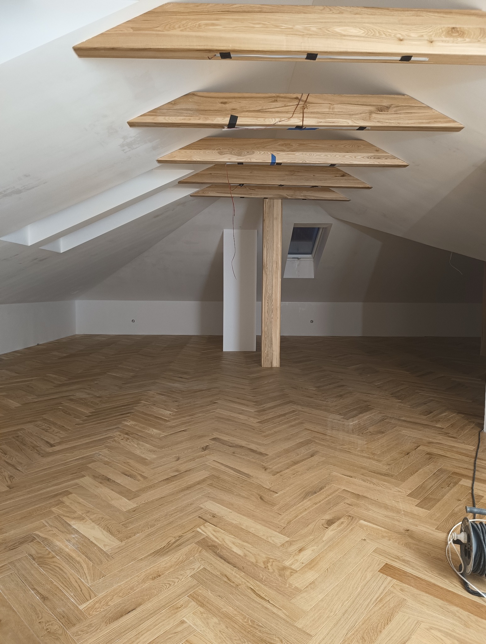 Poddasze z drewnianą podłogą ułożoną w jodełkę, drewnianymi belkami pod sufitem i ścianami pomalowanymi na biało, widoczne przewody elektryczne.