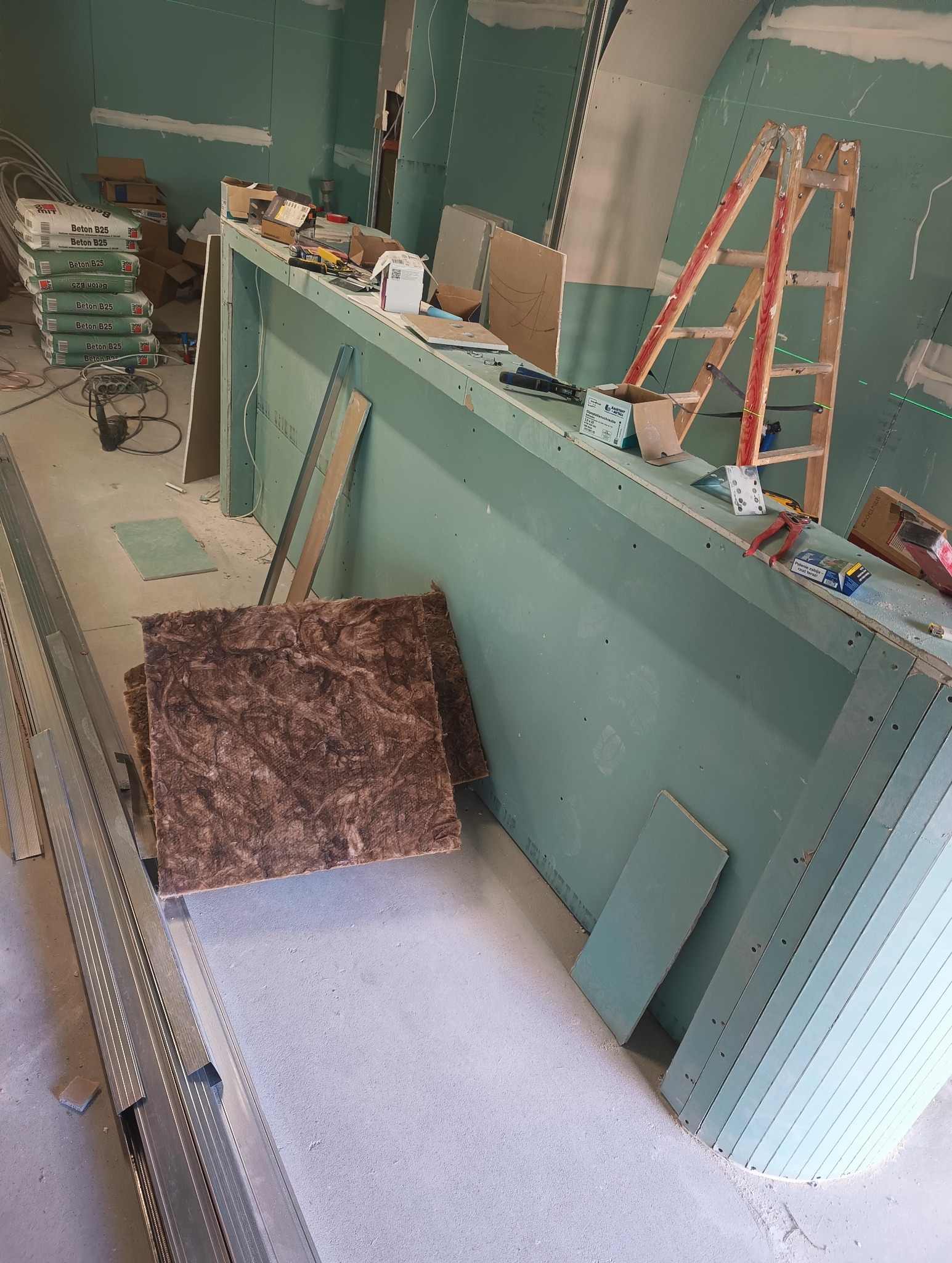 Wnętrze w trakcie prac wykończeniowych: widoczne płyty gipsowo-kartonowe na ścianach, metalowe profile, worki z zaprawą murarską, drabina i narzędzia, sugerujące montaż konstrukcji i przygotowanie...