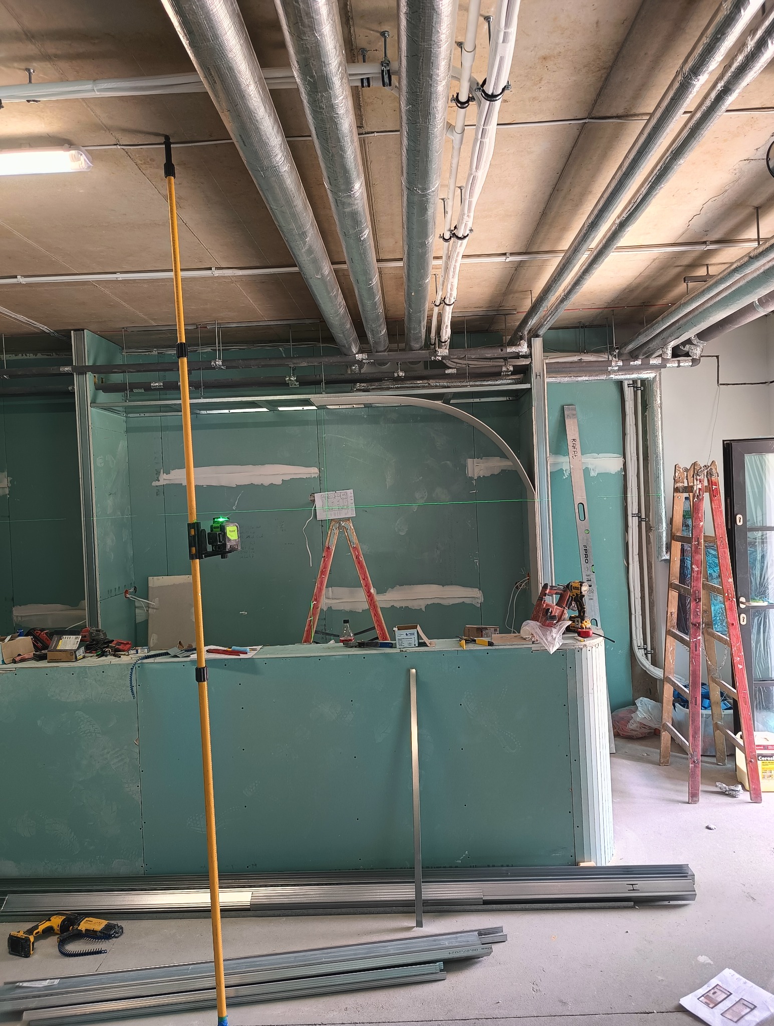 Montaż płyt karton-gips na metalowym stelażu w pomieszczeniu z widocznymi instalacjami pod sufitem, użyciem poziomicy laserowej i drabiny, w trakcie prac wykończeniowych.