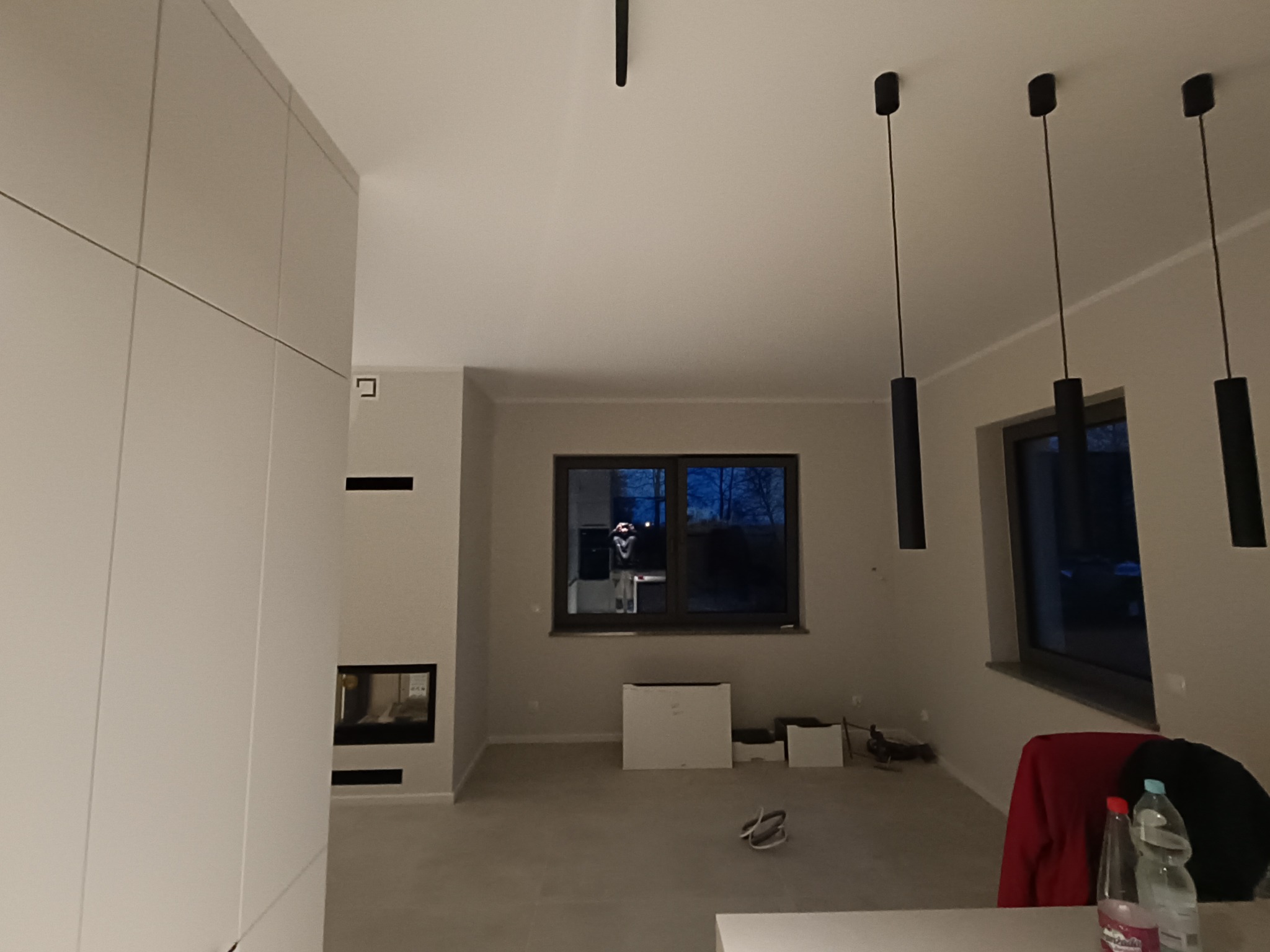 Wnętrze pokoju z minimalistyczną zabudową meblową, kominkiem, trzema wiszącymi lampami i dużym oknem, widoczne ślady po pracach wykończeniowych.