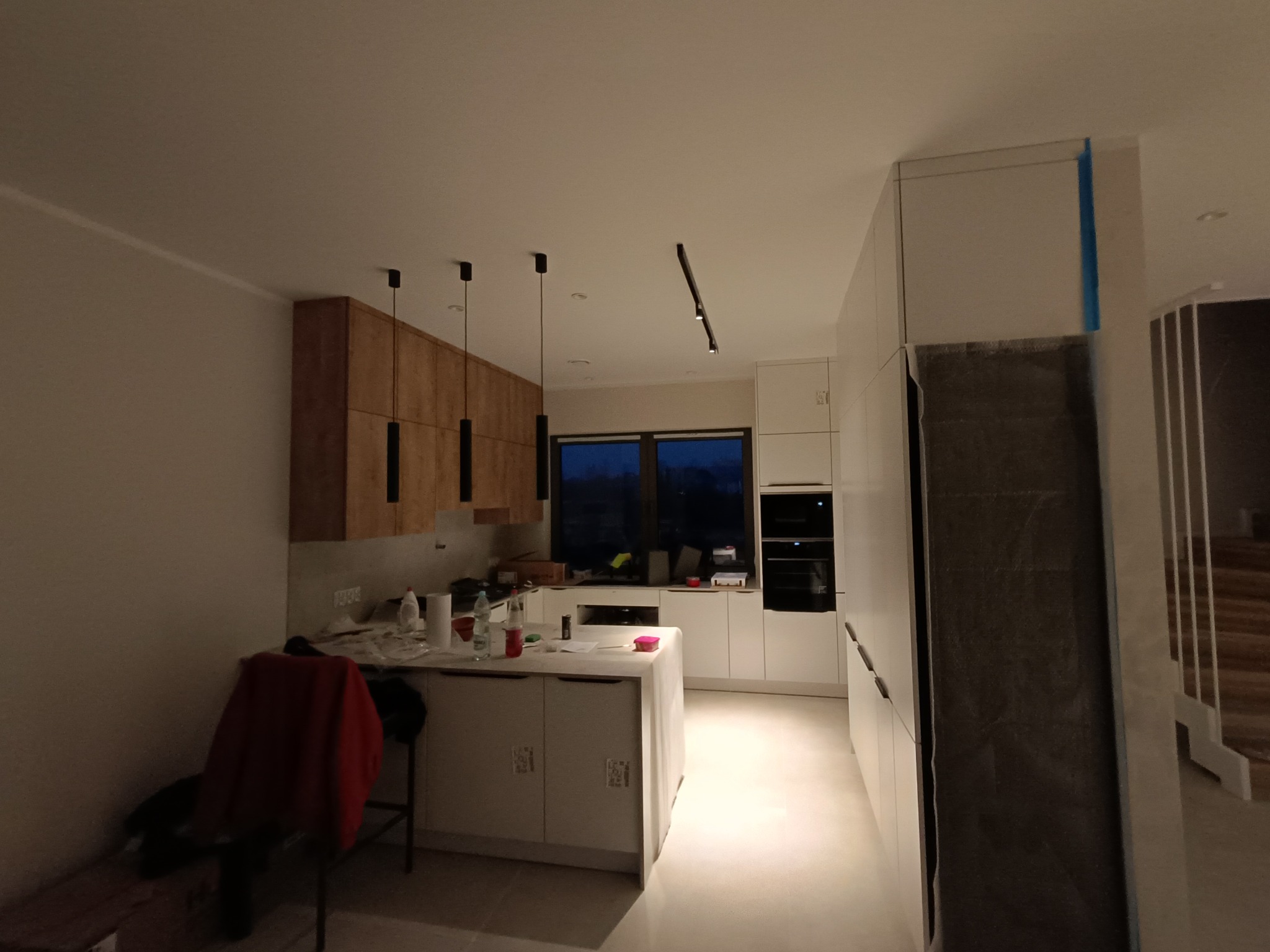 Wnętrze kuchni w trakcie remontu, widoczne drewniane szafki, białe meble kuchenne, wyspa z zabezpieczeniem folią malarską, ściany przygotowane pod malowanie, oświetlenie punktowe.