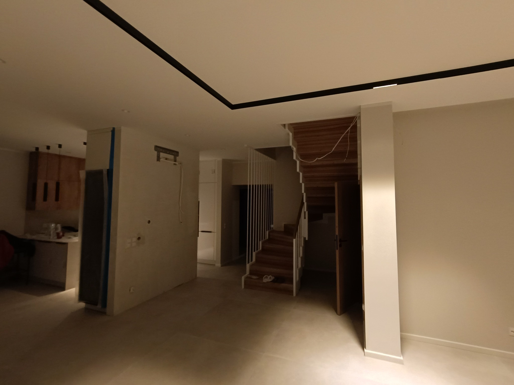 Wnętrze domu w trakcie remontu, widoczne ściany przygotowane do malowania, schody drewniane z białymi barierkami, czarne listwy oświetleniowe na suficie.