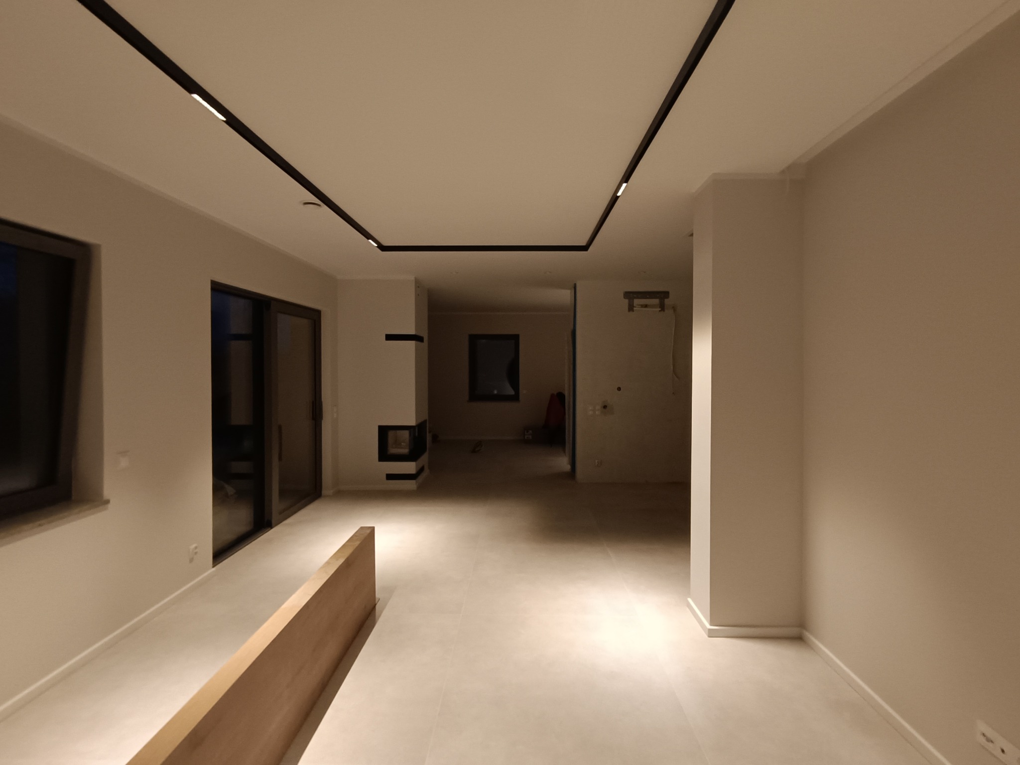 Wnętrze przestronnego, jasnego pomieszczenia z minimalistycznym wykończeniem, oświetleniem szynowym na suficie i kominkiem w oddali.