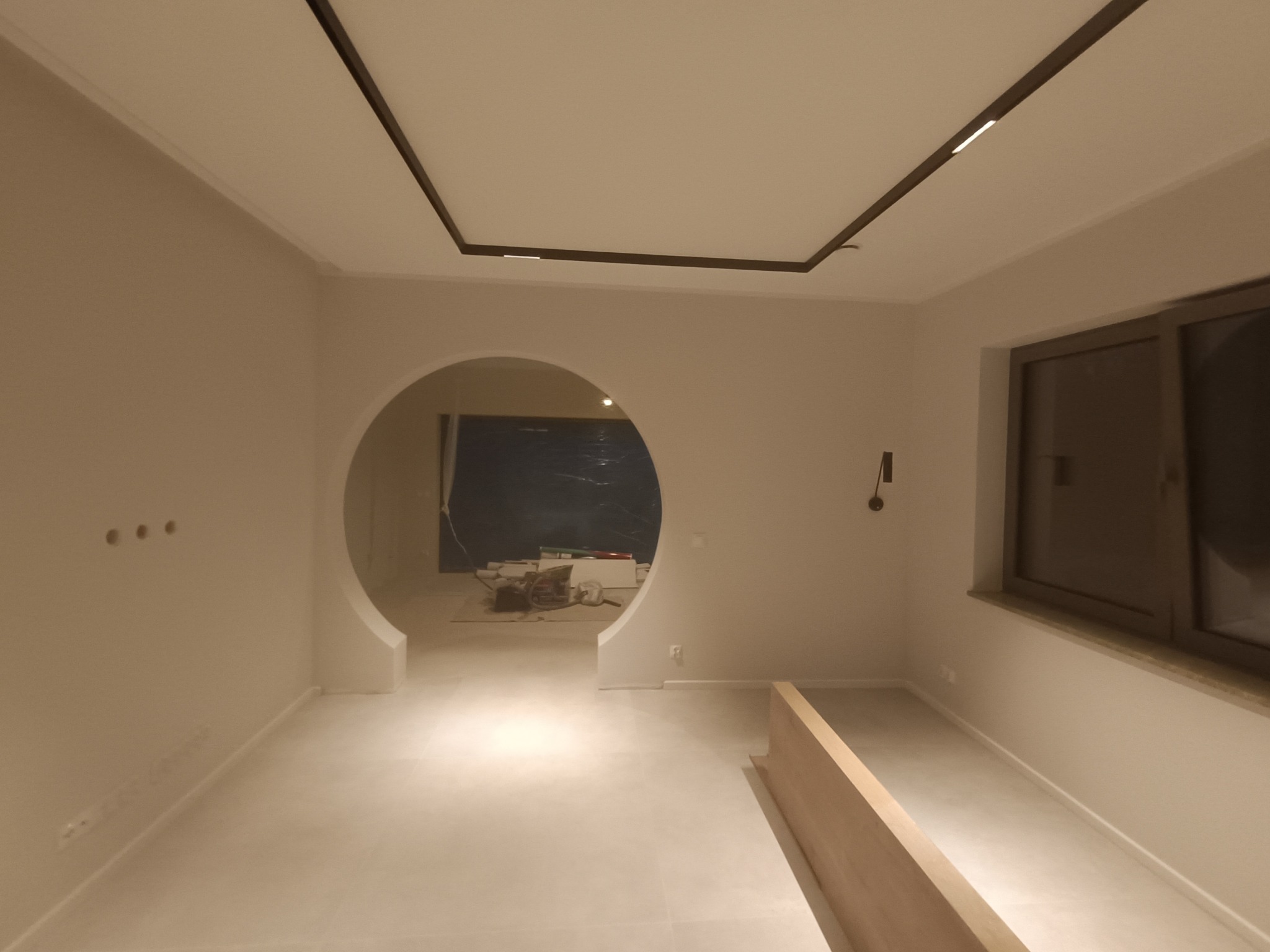 Wnętrze w trakcie wykańczania: białe ściany, podłoga z jasnych płytek, okrągłe przejście do kolejnego pomieszczenia z widocznymi materiałami budowlanymi, nowoczesne oświetlenie sufitowe.