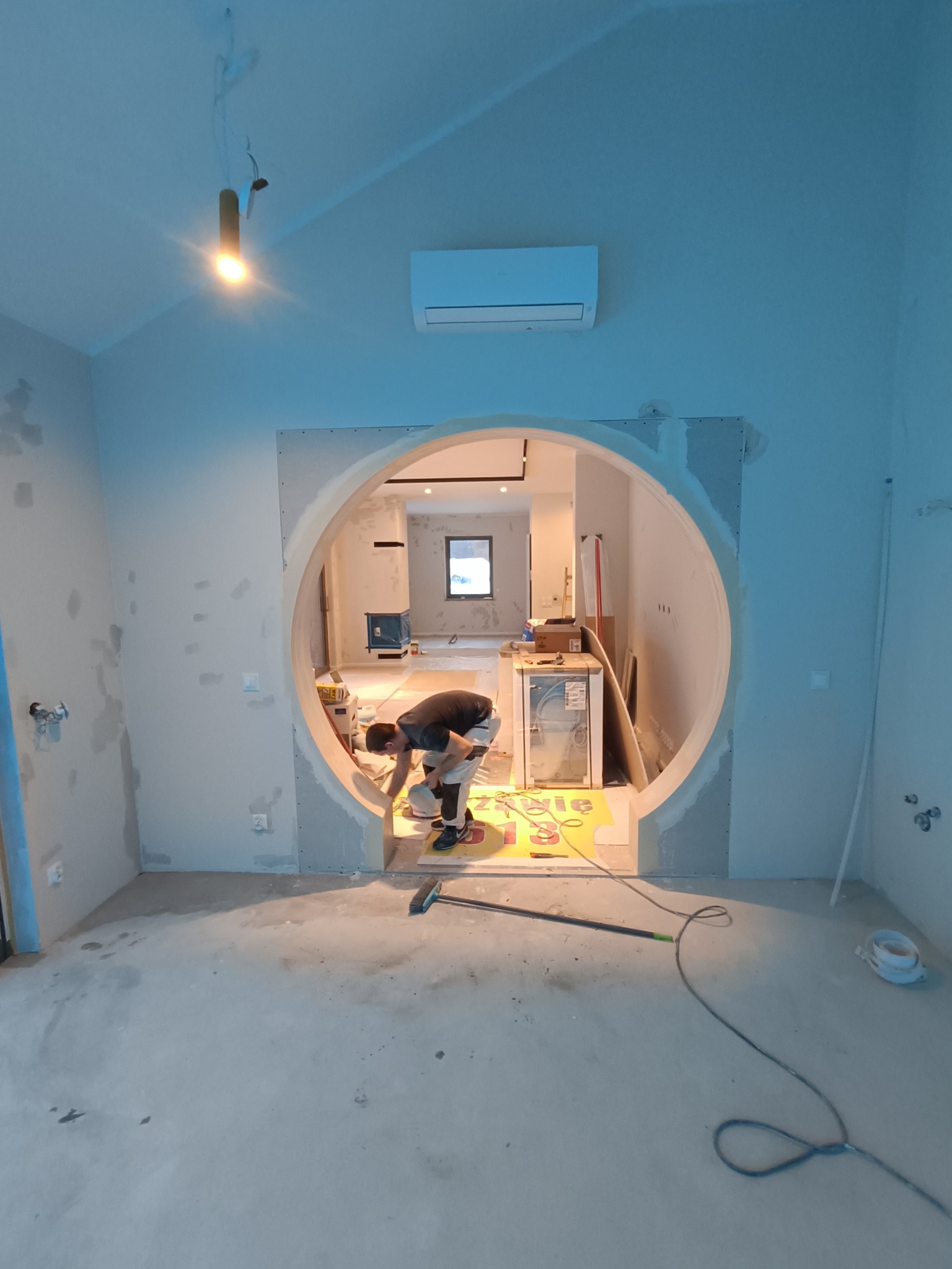 Pracownik w trakcie montażu płyty karton gips na okrągłym otworze w ścianie, widoczne narzędzia, szpachlowane ściany i surowy beton na podłodze w pomieszczeniu z klimatyzacją.