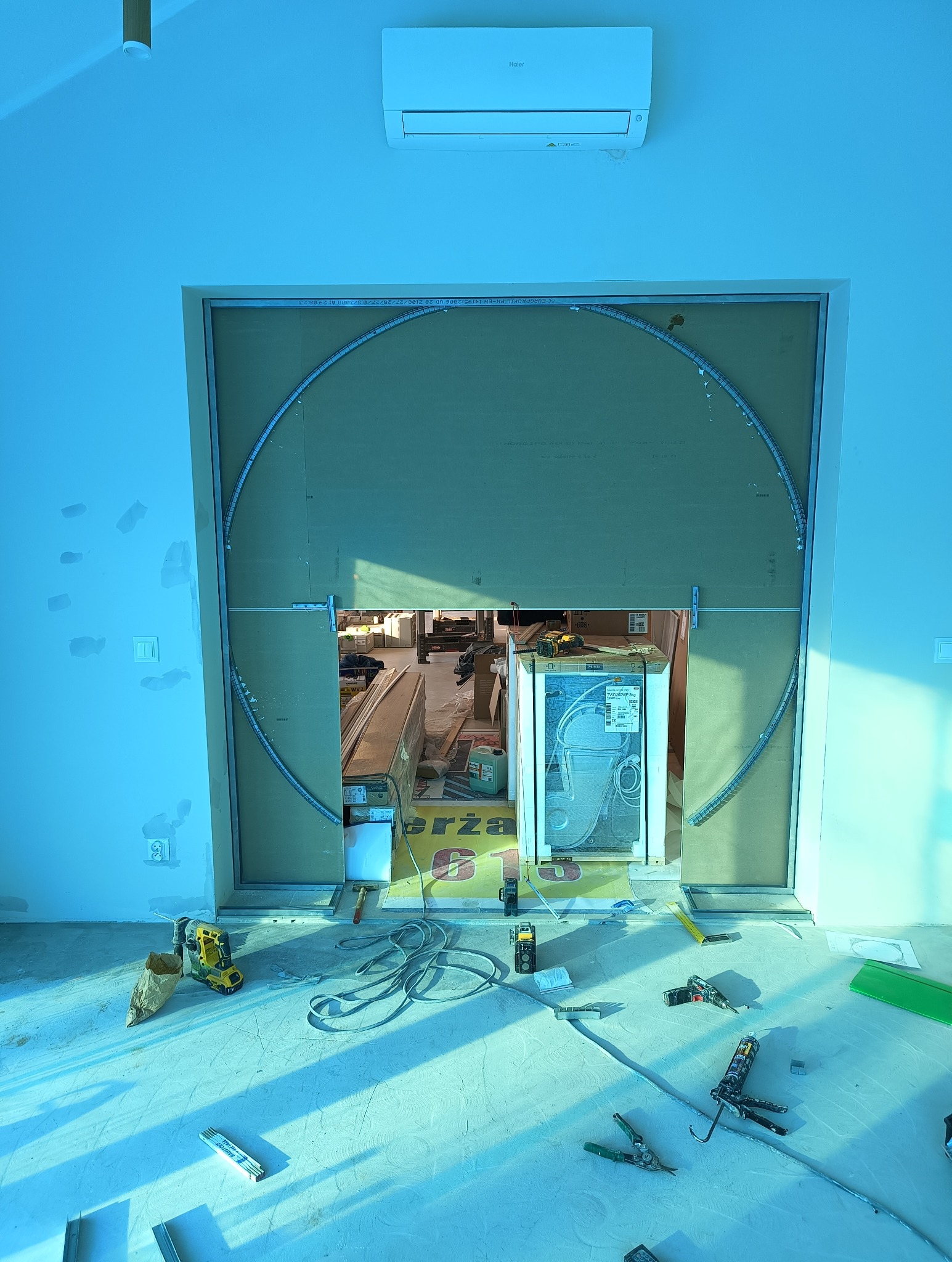 Montaż płyty gipsowo-kartonowej na ścianie z wyciętym okrągłym otworem, widoczne narzędzia i materiały budowlane na podłodze, klimatyzator na górze ściany.