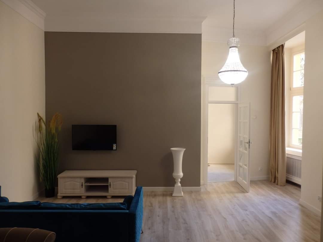 Eleganckie wnętrze po odświeżeniu: ściana w odcieniu taupe z telewizorem, dekoracyjny wazon na postumencie, kryształowy żyrandol i parkiet w jasnym odcieniu dębu.