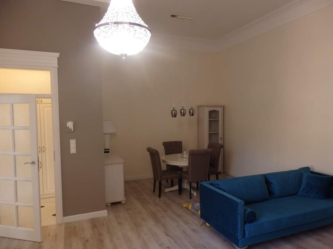 Jasne wnętrze: salon z drewnianą podłogą, niebieską sofą, okrągłym stołem z krzesłami, kryształowym żyrandolem i ścianami w odcieniach beżu i szarości.