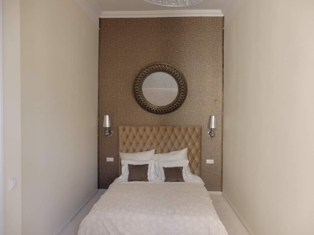 Wąska sypialnia z beżową tapetą, pikowanym zagłówkiem, okrągłym lustrem i symetrycznie rozmieszczonymi kinkietami po bokach. Ściany pomalowane na kremowo.