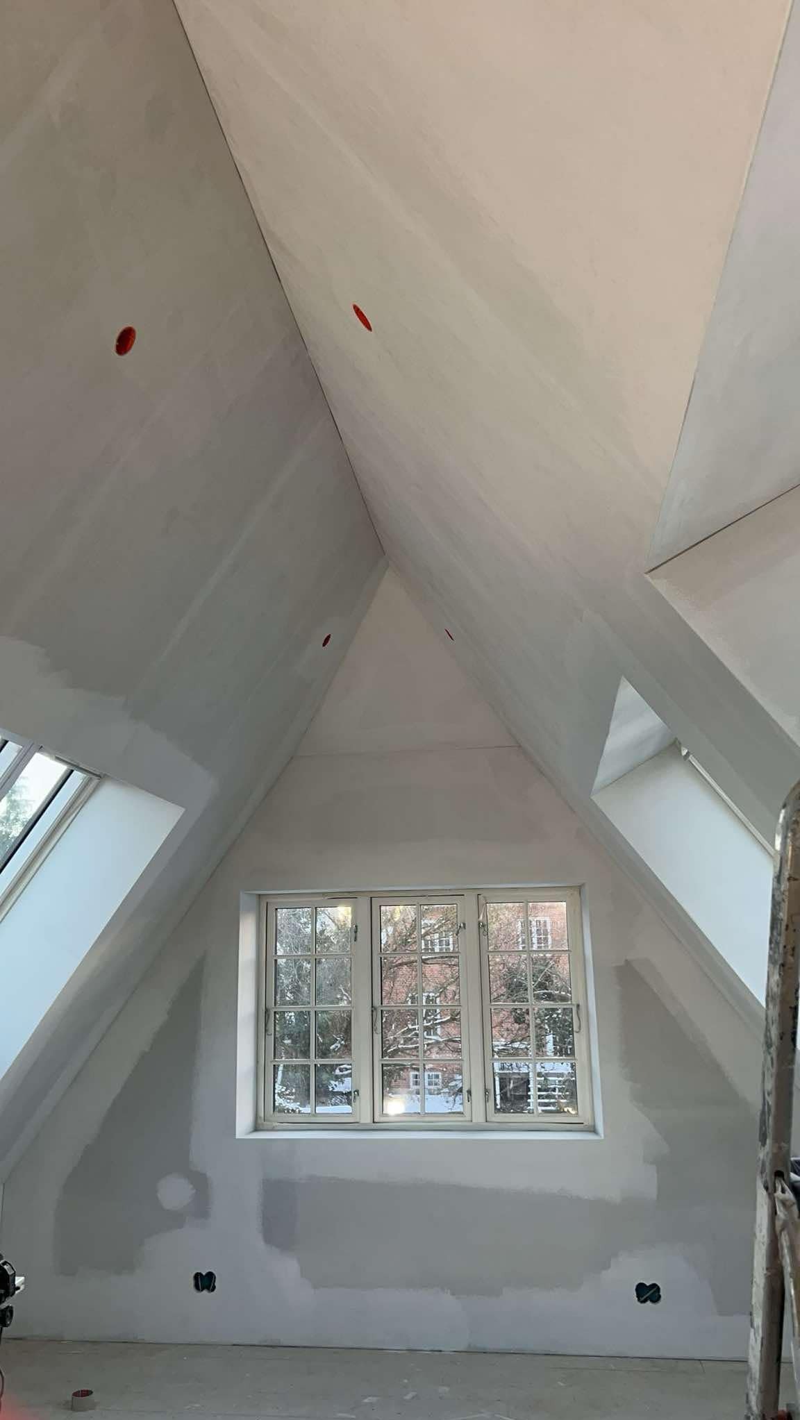 Poddasze ze skosami w trakcie szpachlowania, widoczne okna dachowe i centralne okno z widokiem na budynek z czerwonej cegły, zamaskowane otwory w suficie, ślady szpachli na ścianach.