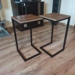 Dwa stoliki kawowe z drewnianymi blatami i czarnymi metalowymi nogami w kształcie litery U, na tle laminowanej podłogi, z widocznym drapakiem dla kota i szafką RTV w tle.