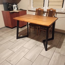 METAL FUSION - Stół z drewnianym blatem i czarnymi, metalowymi nogami o nietypowym kształcie, ustawiony w pomieszczeniu z jasną podłogą imitującą drewno i dwoma krzesłami w tle.