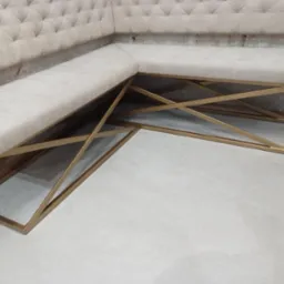 Narożna ławka tapicerowana z pikowanym oparciem, wsparta na geometrycznej konstrukcji z profili w kolorze złotym, ustawiona na jasnej podłodze.